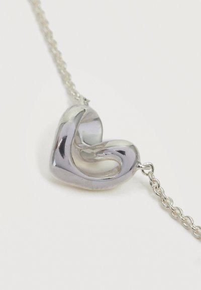 Pendentif en argent incurvé ressemblant à une boucle torsadée sur une délicate chaîne en argent, sur un fond blanc uni.