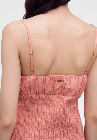 Dos nu d'une femme aux cheveux foncés portant une robe rose froncée avec des bretelles fines réglables et un petit détail de logo doré.