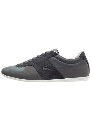 Baskets basses gris foncé avec des panneaux en cuir et daim mélangés, fermeture à lacets, semelle blanche et petit logo crocodile sur le côté.