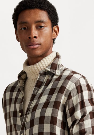 Camisa de franela a cuadros en tonos marrones y crema con cuello camisero, sobre un suéter de cuello alto en crema con relieve.