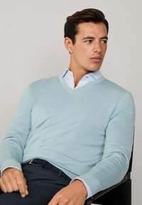 Svetlo modrý sveter s výstrihom do V z jemného materiálu, nosený cez bielo-modrú pruhovanú košeľu s golierom, v kombinácii s tmavými nohavicami a čiernou stoličkou.