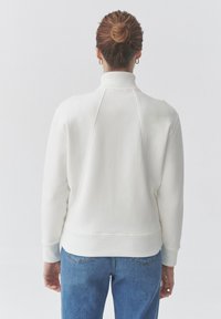 Felpa bianca con collo alto e polsini a coste. Presenta una vestibilità rilassata e dettagli di cucitura visibili sul retro. Indossata con jeans blu.