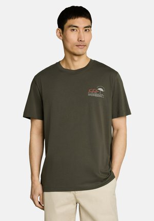 Man draagt een olijfgroene korte mouw T-shirt met een kleine witte en rode afbeelding op de borst, gecombineerd met beige broek, staand tegen een witte achtergrond.