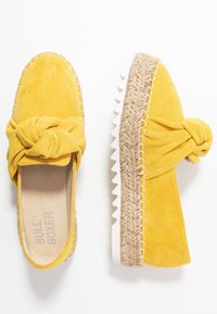 Zapatos amarillos de ante tipo slip-on con detalle de lazo anudado, suela de plataforma de yute y suela exterior de goma blanca con estrías, mostrados desde vistas superiores y laterales.