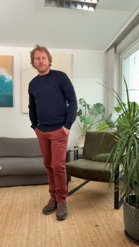Pull bleu foncé, pantalon bordeaux, chaussures grises. Debout dans une pièce avec des plantes, un canapé gris et une chaise verte sur un fond clair.