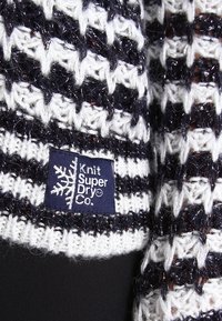 Pull en maille grosse dans les tons noir et blanc, présentant un motif texturé à tissage ouvert. Comprend une étiquette bleue distincte avec l'inscription "Knit Super Dry Co."