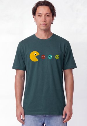 Homme portant un t-shirt vert foncé avec un personnage Pac-Man jaune et trois petits visages colorés en rangée sur le devant.