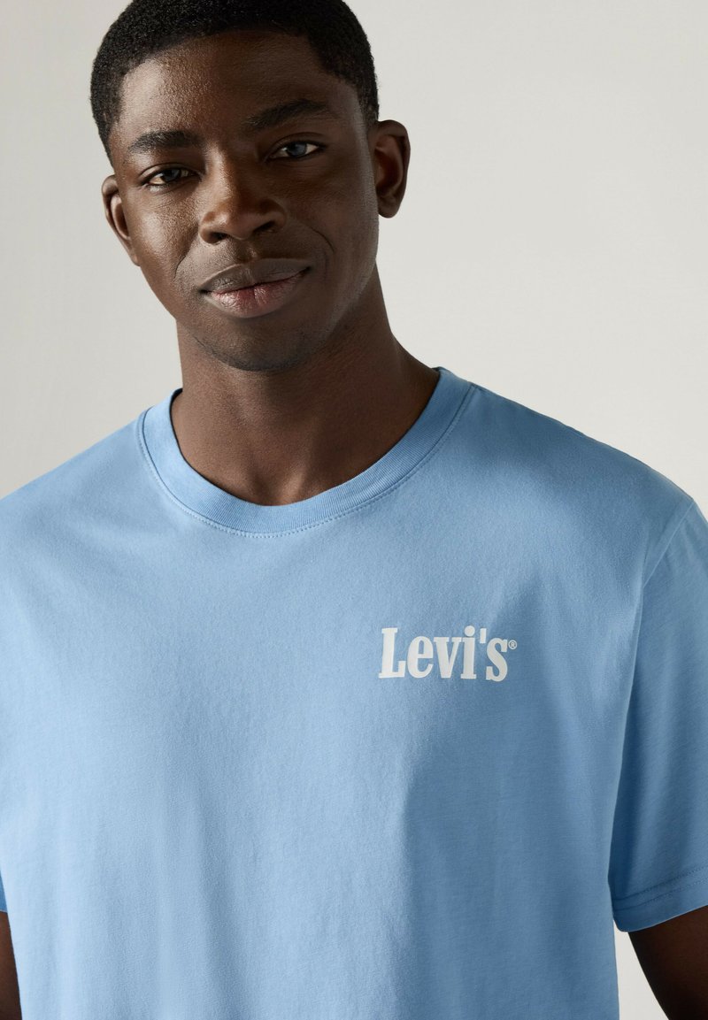 Junger Mann trägt ein hellblaues Levi's T-Shirt und schaut mit neutralem Gesichtsausdruck in die Kamera vor einem einfarbigen Hintergrund.