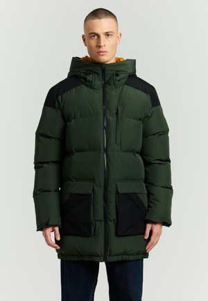 Mężczyzna w zielono-czarnej pikowanej zimowej kurtce z wysokim kołnierzem i kapturem, stojący na jednolitym jasnym tle.