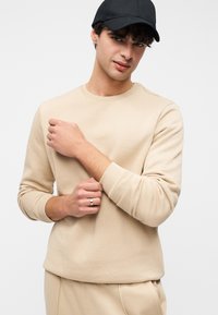 Μπεζ φούτερ με μακριά μανίκια, στρογγυλή λαιμόκοψη και ριμπ μανσέτες. Φορώντας μαύρο καπέλο, το μοντέλο ποζάρει με ένα χέρι στο στήθος.