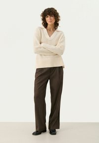 Maglione beige chiaro lavorato a maglia con scollo a V e polsini a costine, abbinato a pantaloni larghi marrone scuro e scarpe nere.