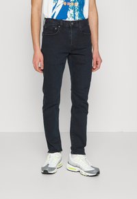 Jeans de mezclilla oscuros ajustados con una textura suave, cinco bolsillos y cierre de cremallera. Combinados con zapatillas deportivas grises y blancas con acentos neón.