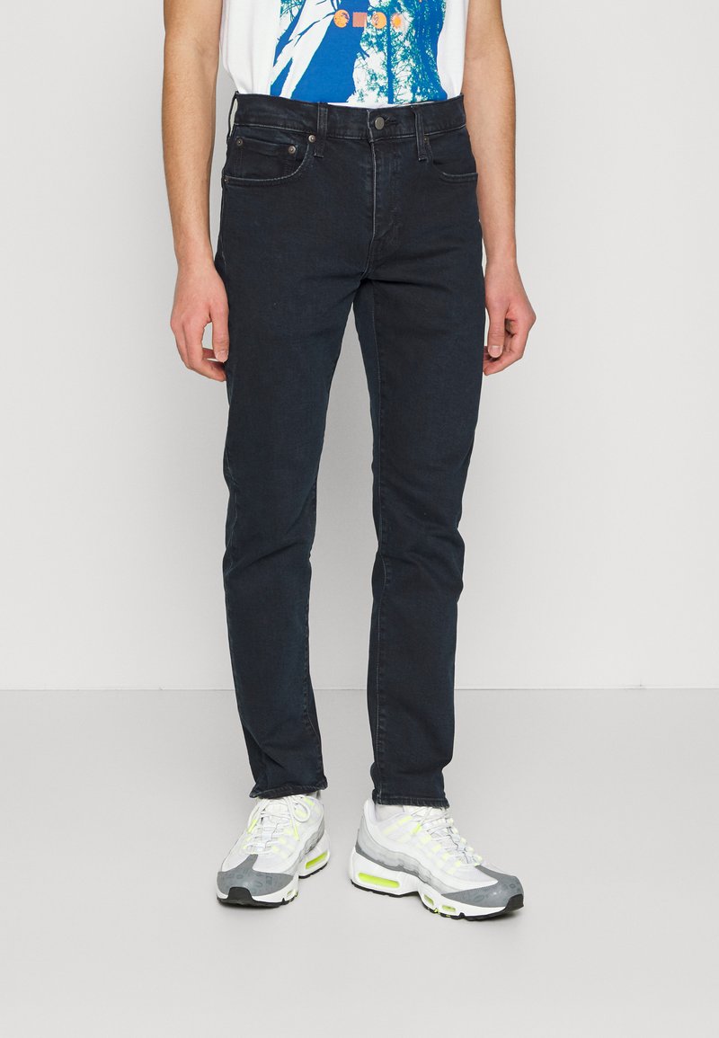 Jeans de mezclilla oscuros ajustados con una textura suave, cinco bolsillos y cierre de cremallera. Combinados con zapatillas deportivas grises y blancas con acentos neón.