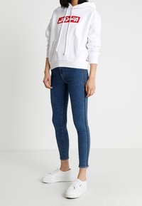 Felpa bianca con cappuccio e logo rosso, abbinata a jeans skinny blu con striscia laterale, e sneakers bianche. Design semplice e vestibilità casual.