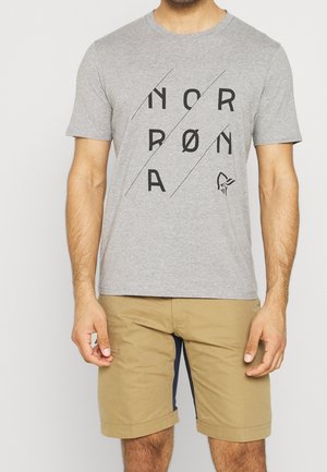 Mężczyzna w jasnoszarej koszulce z napisem "NORRØNA" i logo w postaci hełmu wikinga, ubrany w beżowe szorty.
