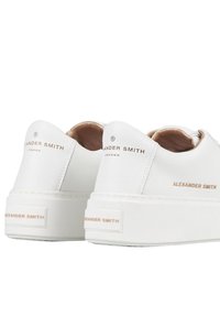 Sneaker bianche in pelle con texture liscia, dotate di una suola spessa bianca e un discreto marchio dorato sul tallone e sulla parte superiore.