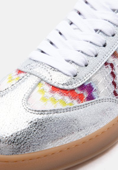 Gros plan sur une basket argentée texturée avec des lacets blancs, un détail de couture en zigzag arc-en-ciel et une semelle marron clair.
