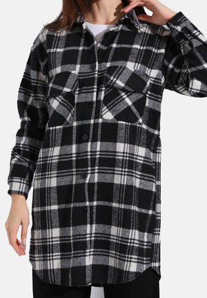 Femme portant une chemise à carreaux noire et blanche à manches longues avec poches poitrine par-dessus un top blanc, ajustant ses cheveux d'une main.