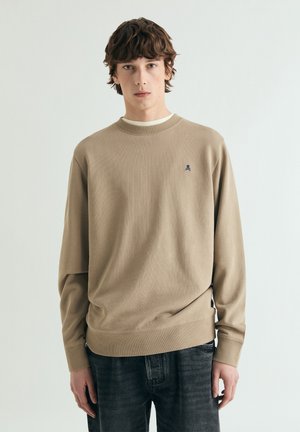 Joven con cabello castaño de longitud media, que lleva una sudadera beige con un pequeño logo de calavera y jeans gris oscuro, posando frente a un fondo blanco.