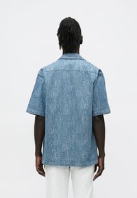 Világoskék rövid ujjú farmering texturált felülettel, gallérral és egyenes alsó szegéllyel. Hátsó nézeten laza szabás és egyszerűség látható.