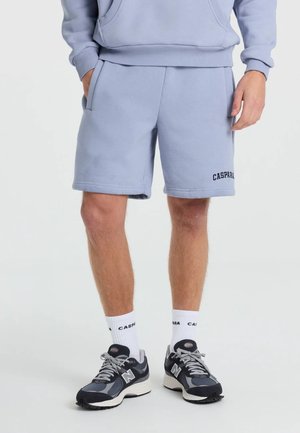 Mand iført lyseblå sweatshorts med sidelommer, hvide mærkesokker og sorte og grå New Balance sneakers, stående med hænderne i lommerne.