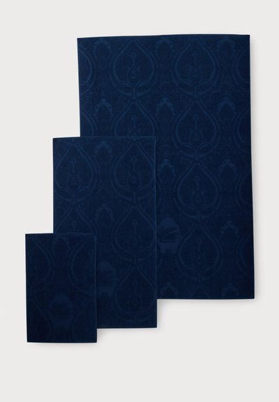 Trois livres en velours bleu foncé empilés avec un subtil motif floral damassé sur leurs couvertures sur un fond clair.