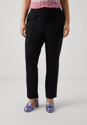 Osoba nosząca czarne spodnie o prostych nogawkach z elastycznym pasem i metaliczne fioletowe buty na szerokim obcasie na jasnym tle.