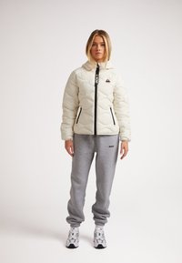 Puffig, ljusfärgad jacka med svart dragkedja och logotyp, tillsammans med gråa träningsoverallsbyxor. Modellen bär vita och gråa sneakers.