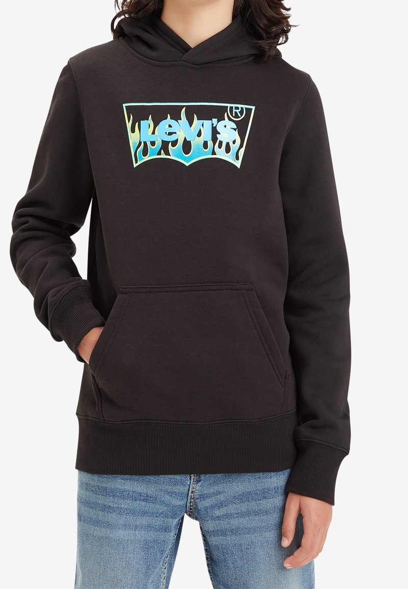 Personne portant un sweat à capuche noir avec un logo Levi's en forme de flamme bleue, une poche avant, et un jean bleu, main dans la poche, sur un fond blanc.