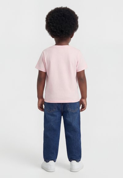 Barn med krøllet hår står med fronten væk, iført en lyserød T-shirt, blå jeans og hvide sneakers på en ensfarvet baggrund.