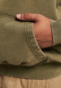 Sweatshirt vert olive avec des poignets côtelés, affichant un tissu doux et texturé. Une main repose dans une poche latérale, mettant en avant le design du vêtement.