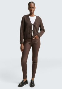 Ženska stoji oblečena v rjavem kardiganu, zavezana z dvema pentljama, beli majici, rjavih skinny hlačah in črnih loaferjih proti enostavni ozadju.