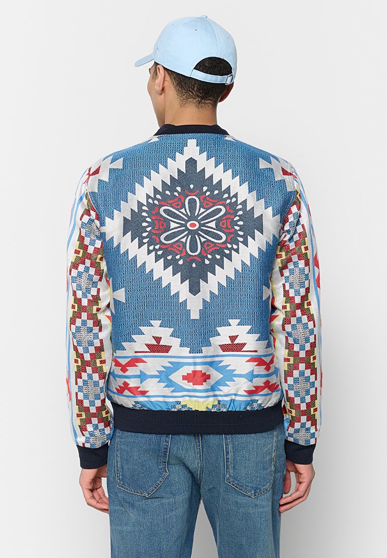 Man in een lichtblauwe pet en een kleurrijke Azteekse patroonjack, staand met zijn rug naar de camera tegen een effen witte achtergrond.