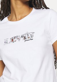 Camiseta blanca con el logotipo colorido de "Levi's" que presenta elementos de la naturaleza y el texto "Un Estado de Sueño", llevada por una persona con cabello trenzado.