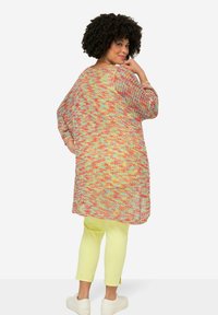 Gebreide cardigan in een meerkleurig patroon met oranje, roze en teal. Losse pasvorm, middellange lengte, gecombineerd met gele cropped broek en witte sneakers.