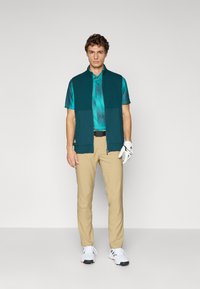 Man stands wearing teal polo shirt, dark green sleeveless vest, beige pants, white golf glove, and white golf shoes on plain background.