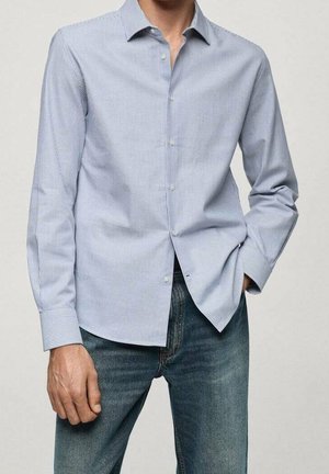 Homme portant une chemise bleu clair à fines rayures et un jean bleu, une main dans la poche, montré de l’épaule à la cuisse.