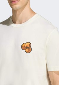 Krémové tričko s grafikou troch zlatých mincí na hrudi. Mäkká látka s klasickým okrúhlym výstrihom a krátkymi rukávmi.