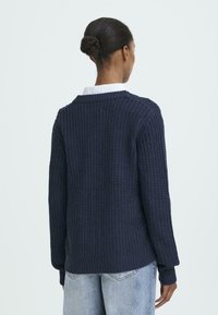 Marinblå stickad tröja med texturerat tyg, ribbade ärmslut och nederkant, med en vid halsringning och avslappnad passform, tillsammans med ljusa denimjeans.