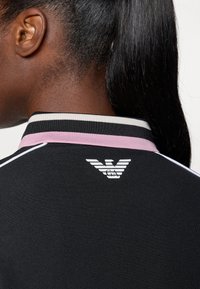 Svart polo skjorta med en krage i rosa och vitt randigt mönster. Vit logotyp på baksidan, minimal struktur och sportig design.