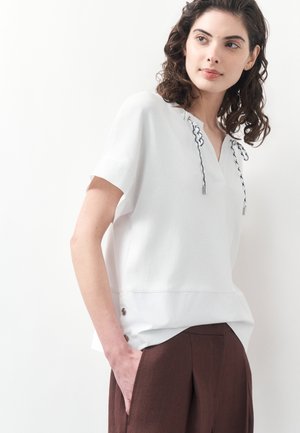 Frau mit lockigem dunklem Haar, die eine weiße kurzärmelige Bluse mit schwarzen Krawattendetails und braune hoch taillierte Hosen trägt, steht mit der Hand in der Tasche.