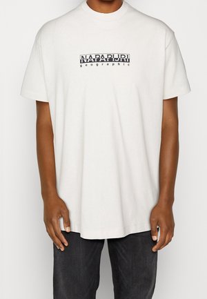 Henkilö, jolla on valkoinen lyhythihainen t-paita, jossa musta "Napapijri geographic" -logo rinnassa, ja tummat housut, yksivärisellä taustalla.