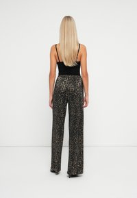 Femeie cu păr lung blond, purtând un top negru fără mâneci și pantaloni largi cu imprimeu de leopard, stând în picioare împotriva unui zid alb simplu.