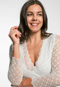 Weißes Spitzen-Wickeloberteil mit tiefem V-Ausschnitt, langen Ärmeln mit gekerbten Kanten und einem strukturierten Diamantmuster.