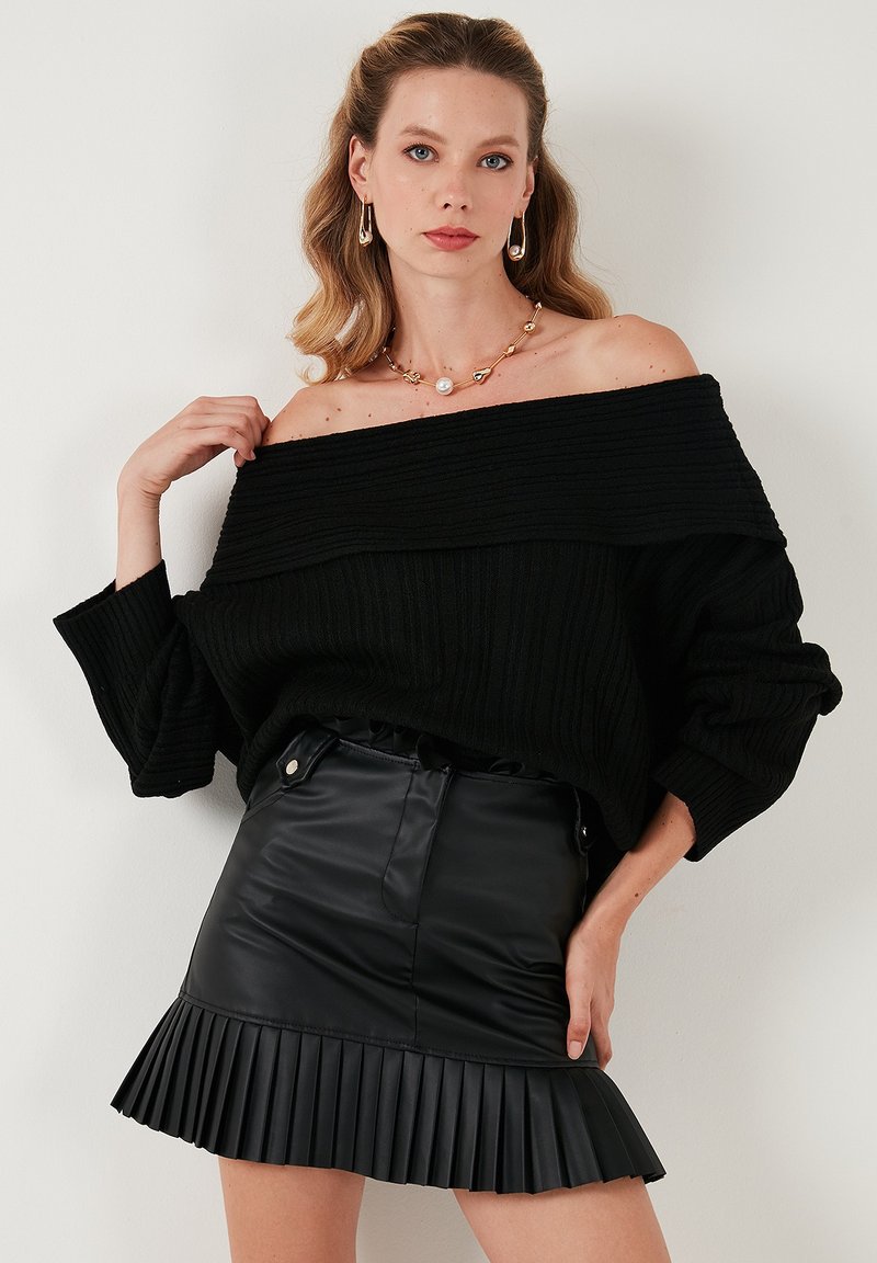 Suéter negro de canalé con los hombros al descubierto, combinado con una falda plisada de cuero sintético negro. La joyería incluye un collar de perlas y oro.