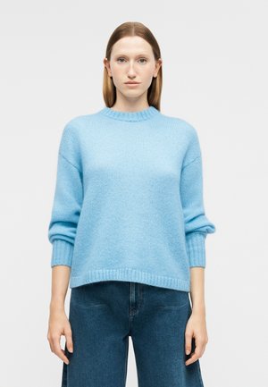Kvinde iført en lyseblå strikket sweater med ribbede ærmekanter og kant forneden, kombineret med højtaljede, mørkeblå denimjeans, stående mod en hvid baggrund.