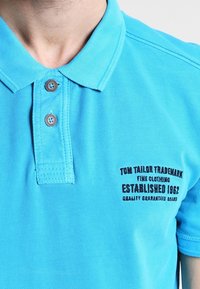 Polo bleu clair en tissu texturé. Présente un col à deux boutons et des détails de logo brodé sur la poitrine gauche.