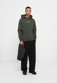 Olivengrøn hoodie med kængurupose og frontlogo, parret med sorte cargo bukser og en quiltet sort taske, stående på en hvid baggrund.