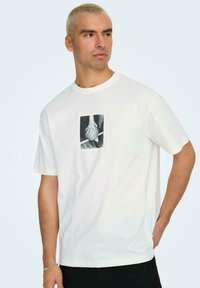 T-shirt in cotone bianco con una fotografia rettangolare in bianco e nero di una mano che tiene un pallone da basket, collo a giro, maniche corte.