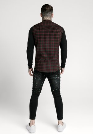 Maglietta nera a maniche lunghe con il retro a quadri bordeaux, abbinata a jeans neri con macchie colorate e sneakers bianche.